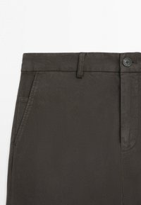 Close-up of dark brown pants waist with belt loops, side pocket, button, and fly against a plain white background.