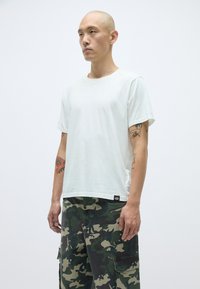 Balts kokvilnas T-kreklis ar īsām piedurknēm un apaļu kakla izgriezumu, apvienots ar zaļiem kamuflāžas biksēm ar kabatām un kontrastējošu šuvumu.