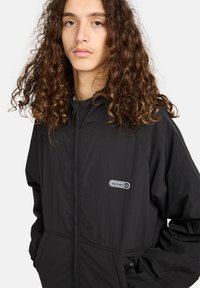 Junge Person mit langen, lockigen braunen Haaren, die eine schwarze Element-Jacke mit Reißverschluss trägt und die Hände in den Taschen hat, vor einem weißen Hintergrund.