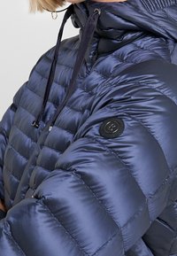 Laivastonsininen toppatakki, jossa tikattu kuviopinta, korkea kaulus, säädettävät kiristysnauhat ja pyöreä logoplaketti hihassa.