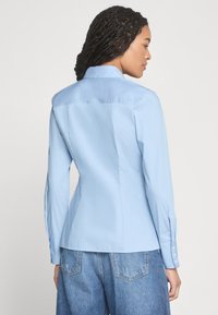 Světle modrá košile s button-up zapínáním, strukturovaným střihem, dlouhými rukávy a zádovým kloboukem. Hladká, tkaná textura s jemnými knoflíkovými akcenty na rukávech.