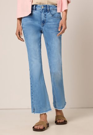 Person iført lyseblå let udvidede jeans med rå kanter og sideslids, kombineret med brune slidersandaler og en lyserød skjorte.