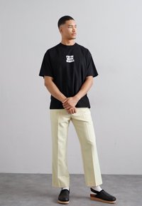 Sort bomulds t-shirt med hvid tekst, cremefarvede bukser og sorte klipklapper med en lysebrun sål. Ensfarvet baggrund med neutrale farver.