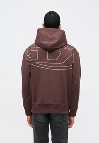 Άνδρας που φοράει ένα σκούρο μωβ φούτερ με αφηρημένο σχέδιο γραμμών στην επάνω πλάτη, συνδυασμένο με σκούρες γ�κρίζες τζιν, γυρισμένος προς τα πίσω.