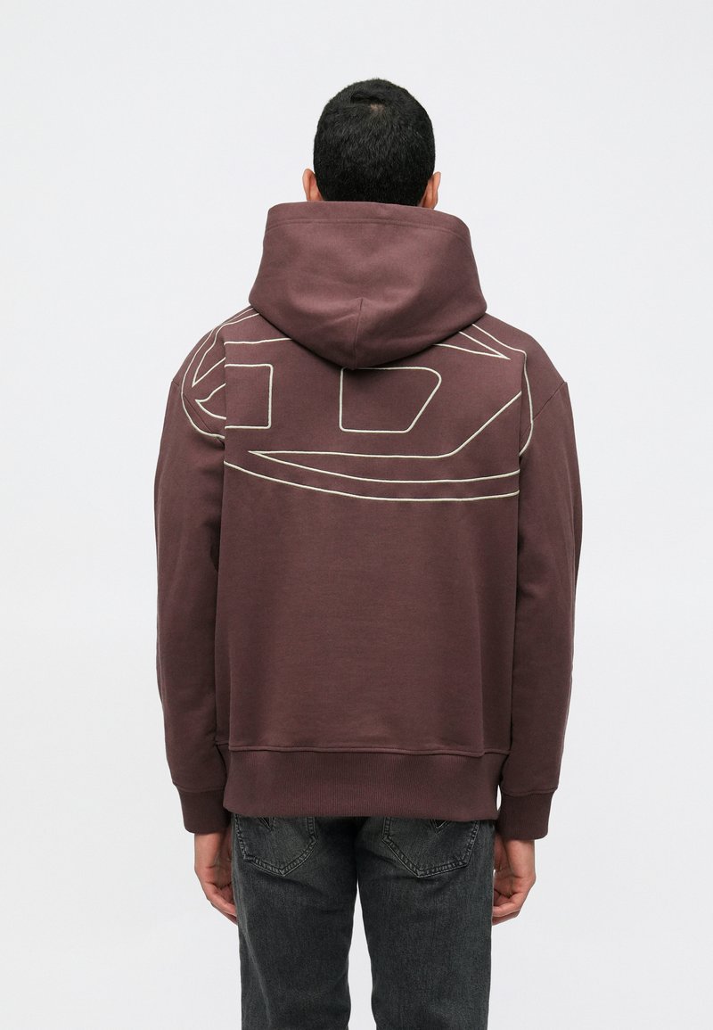 Άνδρας που φοράει ένα σκούρο μωβ φούτερ με αφηρημένο σχέδιο γραμμών στην επάνω πλάτη, συνδυασμένο με σκούρες γκρίζες τζιν, γυρισμένος προς τα πίσω.