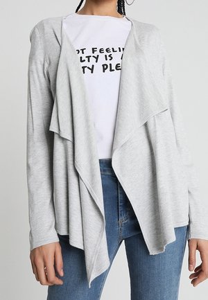 Cardigan ouvert gris clair en tissu doux, avec un design drapé et des manches longues. Porté sur un t-shirt blanc à motif graphique et un jean.