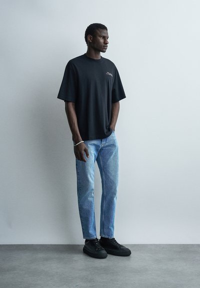 Homme debout contre un mur blanc portant un t-shirt noir surdimensionné, un jean bleu clair, des baskets noires et un bracelet au poignet gauche.