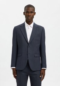 Μπλε σκούρο fitted σακάκι από εκλεπτυσμένο ύφασμα, με κλείσιμο δύο κουμπιών, πέτο με σχίσιμο και δύο μπροστινές τσέπες. Φοριέται πάνω από ένα λευκό πουκάμισο.