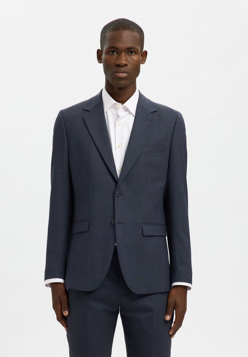 Μπλε σκούρο fitted σακάκι από εκλεπτυσμένο ύφασμα, με κλείσιμο δύο κουμπιών, πέτο με σχίσιμο και δύο μπροστινές τσέπες. Φοριέται πάνω από ένα λευκό πουκάμισο.