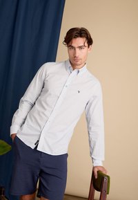 Lichtblauwe gestructureerde overhemd met een button-down kraag, lange mouwen en een borstlogo, gecombineerd met marineblauwe shorts. Detail van een houten stoel zichtbaar.