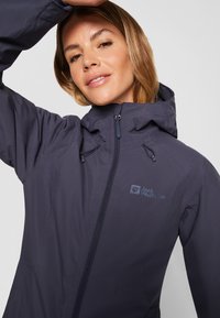 Giacca impermeabile blu navy con cappuccio regolabile, zip frontale completa e logo sul lato sinistro del petto. Texture liscia, design minimalista.