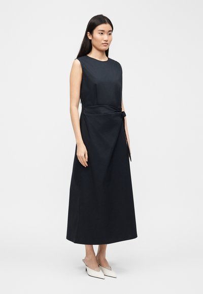 Femme aux longs cheveux noirs, portant une robe midi noire sans manches avec un nœud sur le côté et des escarpins blancs à bout pointu et talons bas, debout devant un fond uni.