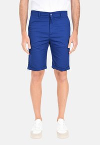 Pantaloni corti in cotone blu con una trama liscia, caratterizzati da un orlo arrotolato e chiusura con bottone frontale, indossati con sneakers bianche.