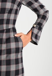 Rutig klänning i svart och vitt med en figurnära överdel, långa ärmar med knapppynt och sidofickor. Strukturerat tyg.
