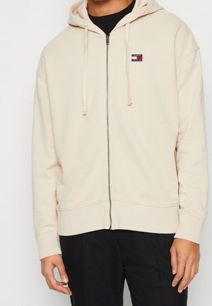 Man draagt een crèmekleurige hoodie met rits en een klein Tommy Jeans-logo op de borst, gecombineerd met een zwarte broek, handen ontspannen langs het lichaam.