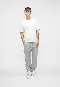 Νεαρός άνδρας που φοράει λευκό μπλουζάκι, γκρι φόρμες και λευκά αθλητικά παπούτσια, με ένα τατουάζ στον αριστερό του καρπό σε απλό λευκό φόντο.