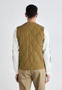 Vesta edidata, de culoare verde măsliniu, prezintă un model în valuri, design fără mâneci și panouri contrastante pe umeri. Purtați-o peste o cămașă albă cu mânecă lungă.