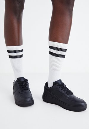 Dark-skinned legs wearing white crew socks with two black stripes and black low-top sneakers standing on a white floor.
