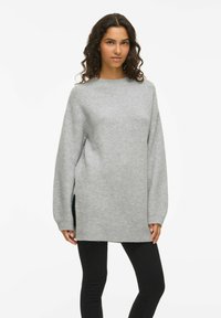 Sieviete ar lokainiem matiem, ģērbusies garā, vaļīgā pelēkā džemperī un melnās piestiprinātajās biksēs, stāv uz baltā fona.