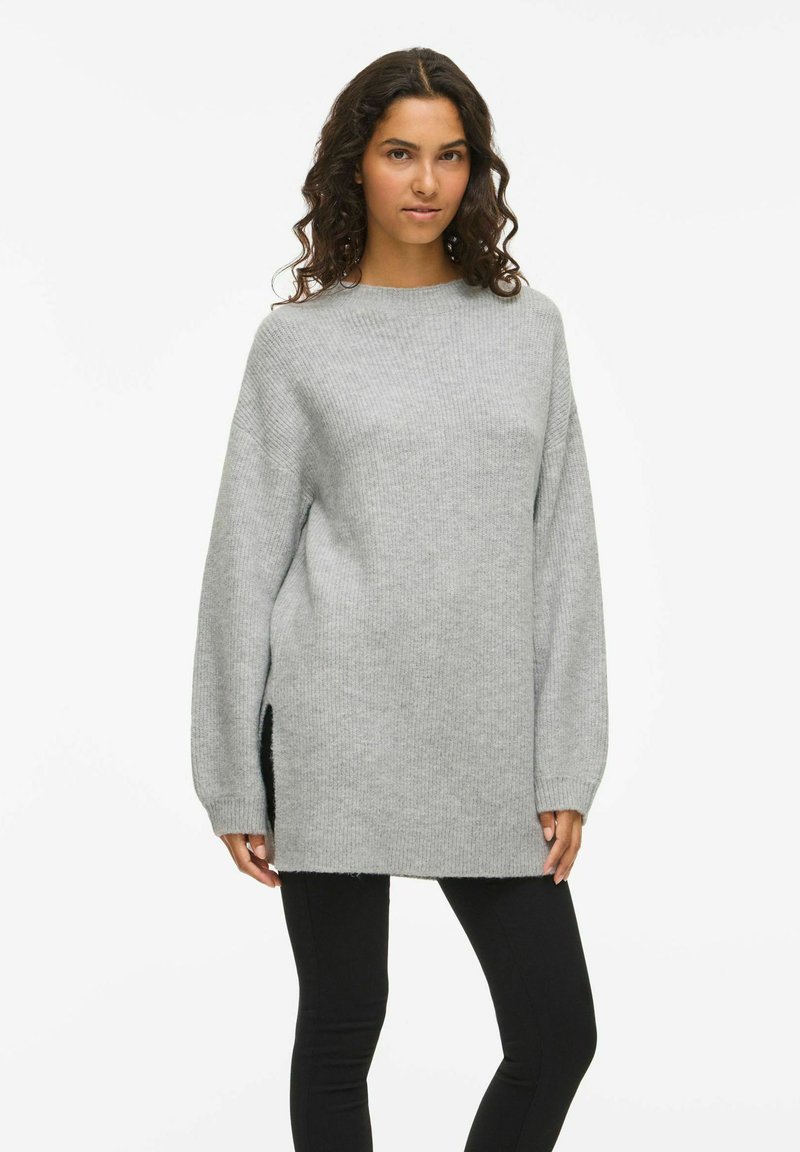 Sieviete ar lokainiem matiem, ģērbusies garā, vaļīgā pelēkā džemperī un melnās piestiprinātajās biksēs, stāv uz baltā fona.