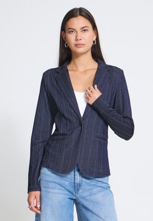 Femme aux longs cheveux foncés portant un blazer cintré foncé à fines rayures sur un haut blanc et un jean bleu clair, debout devant un fond uni.