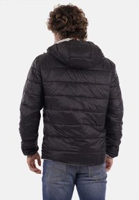 Chaqueta acolchada negra, ligera y con capucha, con un diseño acolchado y una textura suave, usada sobre unos jeans azul. Vista trasera mostrada.