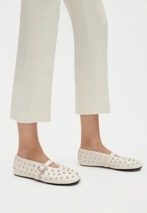 Białe płaskie buty z ozdobami z ćwieków, z zaokrąglonym noskiem i paskiem wokół kostki. Materiał wydaje się gładki o matowym wykończeniu.