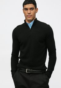 Vyras dėvi juodą megztinį su ketvirčiu užtrauktuku, po juo yra mėlyna marškinė su apykakle, juodus kelnes ir juodą diržą, stovi su rankomis kišenėse.