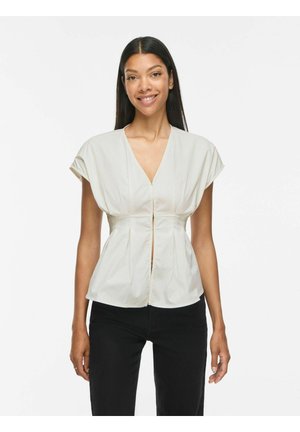 Femme aux longs cheveux foncés portant un chemisier blanc à manches courtes et un pantalon noir, souriante sur un fond blanc uni.