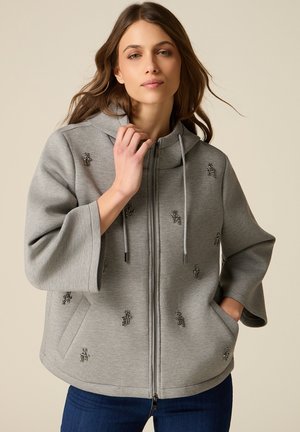 Kobieta nosi jasnoszarą bluzę na zamek z szerokimi rękawami i małymi skupiskami koralików, w połączeniu z ciemnoniebieskimi dżinsami, na neutralnym tle.