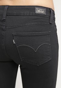 Jeans de mezclilla negra con una vista en primer plano del bolsillo trasero que presenta un patrón de costura curvado distintivo y una etiqueta de marca en la cintura.