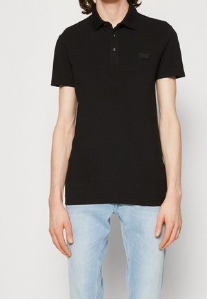 Polo noir avec col, patte de boutonnage à trois boutons et une pièce de marque texturée sur la poitrine. Associé à un jean bleu clair.
