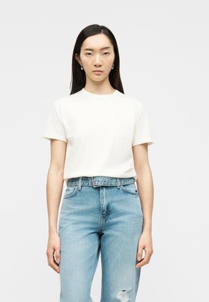 Γυναίκα με μακριά ίσια μαλλιά φοράει απλό λευκό κοντομάνικο μπλουζάκι μέσα σε ψηλόμεσο ανοιχτό μπλε τζιν με σκισμένο γόνατο.