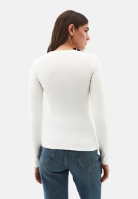 Donna vista di spalle che indossa un top a maniche lunghe bianco attillato e jeans in denim blu su uno sfondo chiaro e uniforme.
