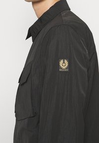 Black lightweight jacket featuring a front pocket, a textured finish, and a prominent Belstaff logo patch on the left shoulder.