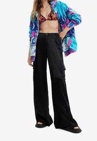 Kolorowa kurtka w kwiaty, czarne spodnie o szerokich nogawkach i sandały na paski. Materiał wydaje się lekki i casualowy, z widocznymi zamkami błyskawicznymi i ozdobnymi guzikami.