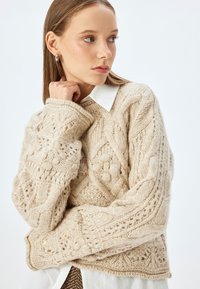 Maglione beige a maglia con motivi intricati, compresi trecce e pompon. Materiale in lana testurizzata e maniche ampie e larghe. Camicia bianca sotto.