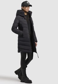 Doudoune noire avec un col montant et une fermeture éclair, dotée de poches latérales, de manches longues et d'un design mi-long. Portée par-dessus des leggings noirs et des bottes.