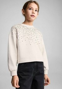 Felpa beige chiaro con scollo rotondo e maniche a palloncino, decorata con abbellimenti scintillanti nella parte superiore anteriore. Abbinata a jeans neri.