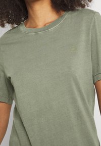 Kortärmad t-shirt med rundad hals i olivgrön bomull, med subtil logotyp och avslappnad passform. Slät yta med minimala sömnadsdetaljer.