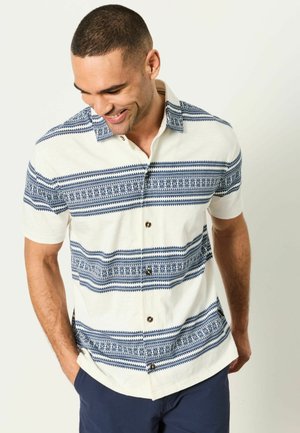 Mann lächelt und schaut nach unten, trägt ein weißes kurzärmeliges Hemd mit marineblauen geometrischen horizontalen Streifen und marineblaue Hose.