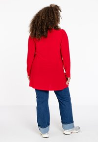 Rood long-sleeve shirt met pofmouwen, gecombineerd met blauwe jeans met opgerolde zomen. Het haar van het model is krullend en schouderlang.