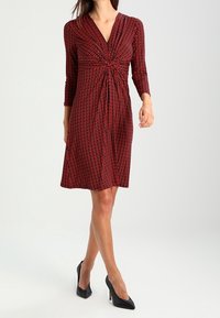 Robe à motifs rouges avec un décolleté en V profond et des manches trois-quarts, dotée d'un détail torsadé à la taille et d'une jupe évasée. Portée avec des talons noirs.