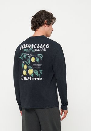 Mand med krøllet hår iført sort langærmet trøje med citrongren-grafik og italiensk tekst på bagsiden, står mod en hvid baggrund.