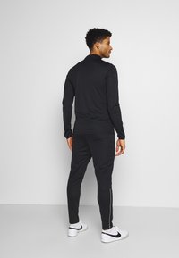 Zwart sportief trainingspak met een longsleeve top en aansluitende broek, vervaardigd uit een gladde stof met witte zijstrepen en subtiele stikseldetails.