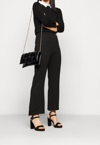 Svart åtsittande tröja, vit skjorta med krage under, svarta vida byxor och svarta sandaler med klack och remmar. Svart vadderad handväska.