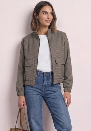 Frau trägt eine braun karierte Jacke, weißes Hemd, blaue Jeans und hält eine beige Handtasche, steht vor einer hellen Wand.