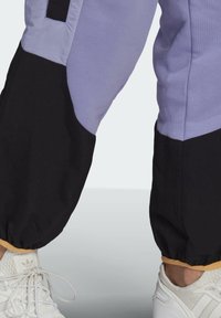 Underben med lila och svarta byxor med elastiska manschetter, ihop med vita Adidas-sneakers mot en ljusgrå bakgrund.