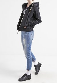 Svart huva jacka med vit dragkedja, kombinerad med ljusblå slitna jeans och svarta sneakers. Modellen står framför en enkel bakgrund.