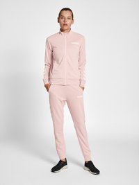 Helles rosa Trainingsanzug aus glattem Material, bestehend aus einer Jacke mit Reißverschluss, die ein weißes Logo und Streifen aufweist, kombiniert mit passenden Jogginghosen und schwarzen Sneakers.